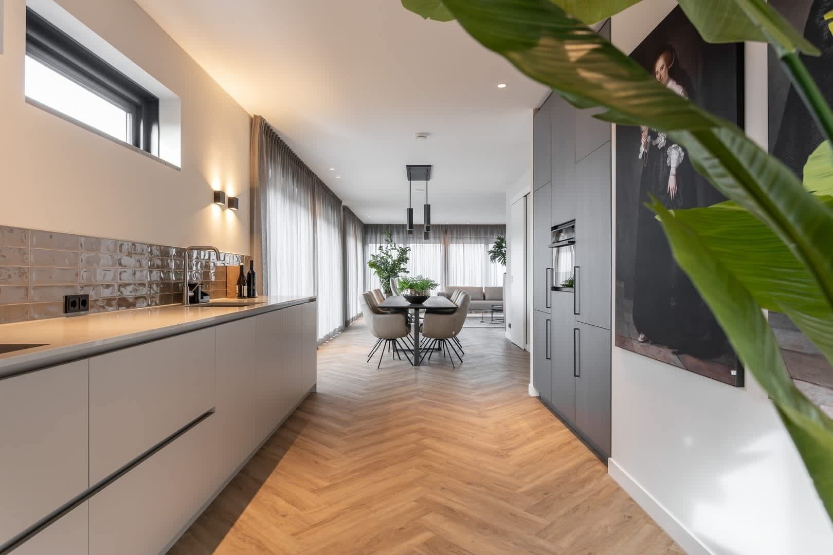 Een moderne keuken met een visgraatvloer, een eetgedeelte met een ronde tafel en stoelen, en een groot raam met gordijnen.