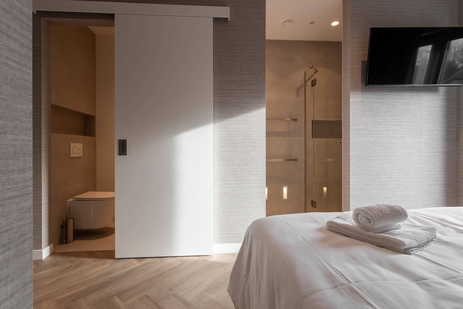 Une chambre d’hôtel moderne avec un plancher en bois, une cabine de douche en verre, un lit avec une serviette roulée et un téléviseur à écran plat fixé au mur.