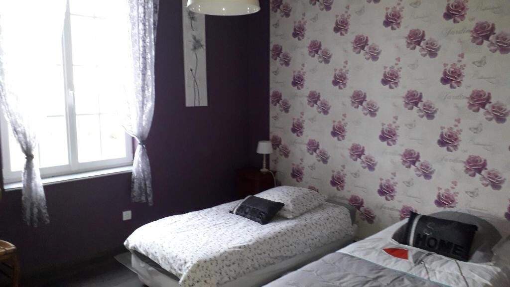 A bedroom with two beds, a window with curtains, a wall with a floral pattern, and a bedside table with a lamp.