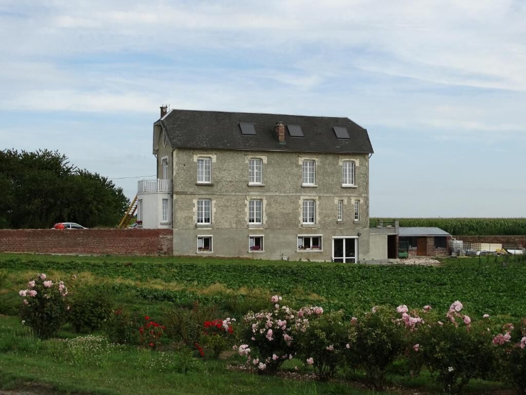 A two-story building with a flat roof and multiple windows, surrounded by a garden with flowering plants.