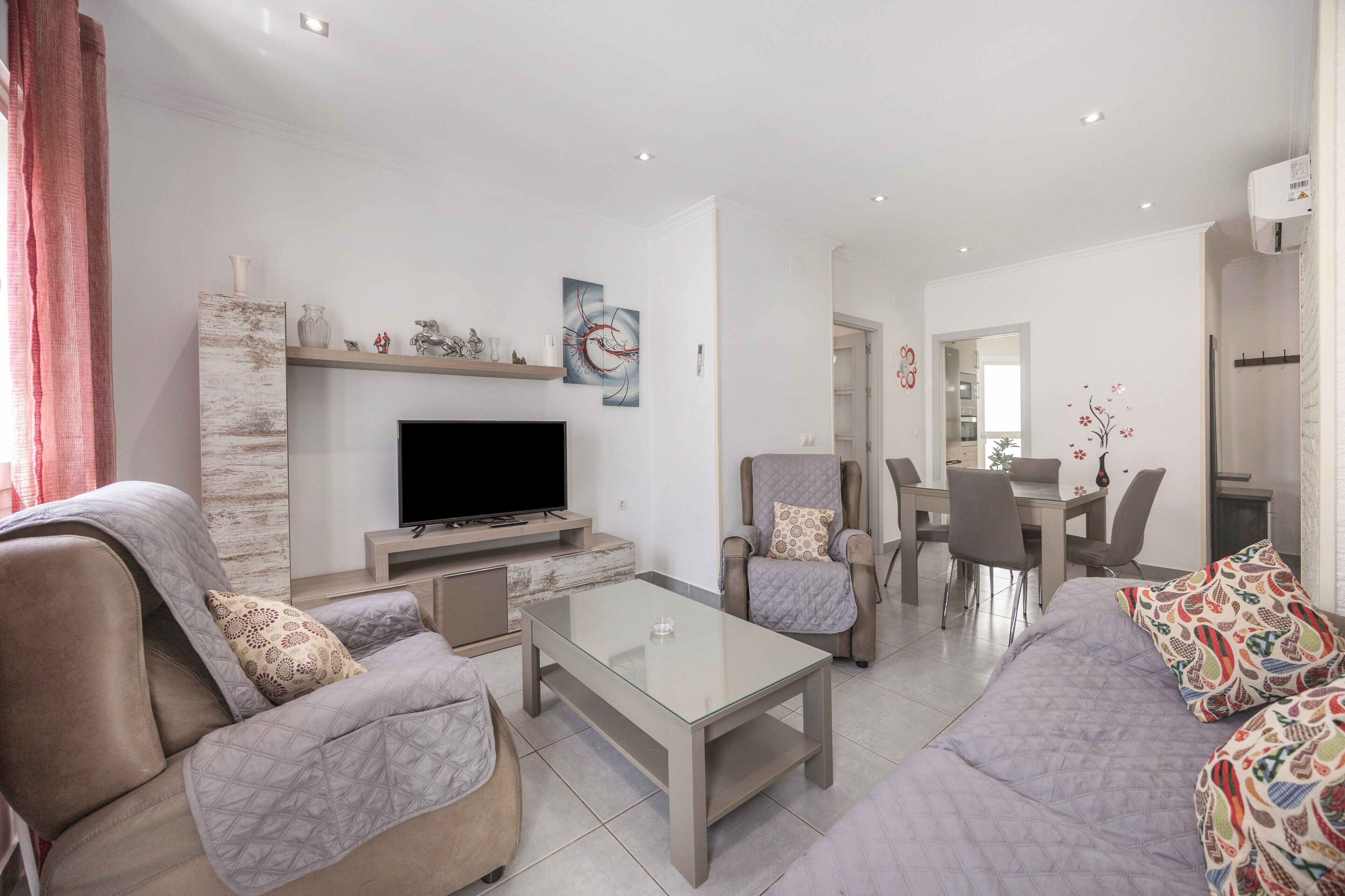 A modern living room with a flat-screen TV, a glass coffee table, sofas, and a dining area with chairs.