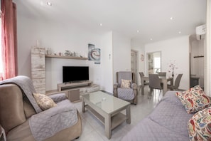 Ein modernes Wohnzimmer mit Flachbildfernseher, einem Glastisch, Sofas und einem Essbereich mit Stühlen.