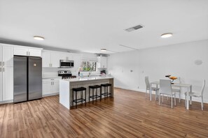 Eine moderne Küche mit weißen Schränken, einem Edelstahlkühlschrank, einer Mikrowelle und einem Essbereich mit weißen Stühlen und einem kleinen Tisch.