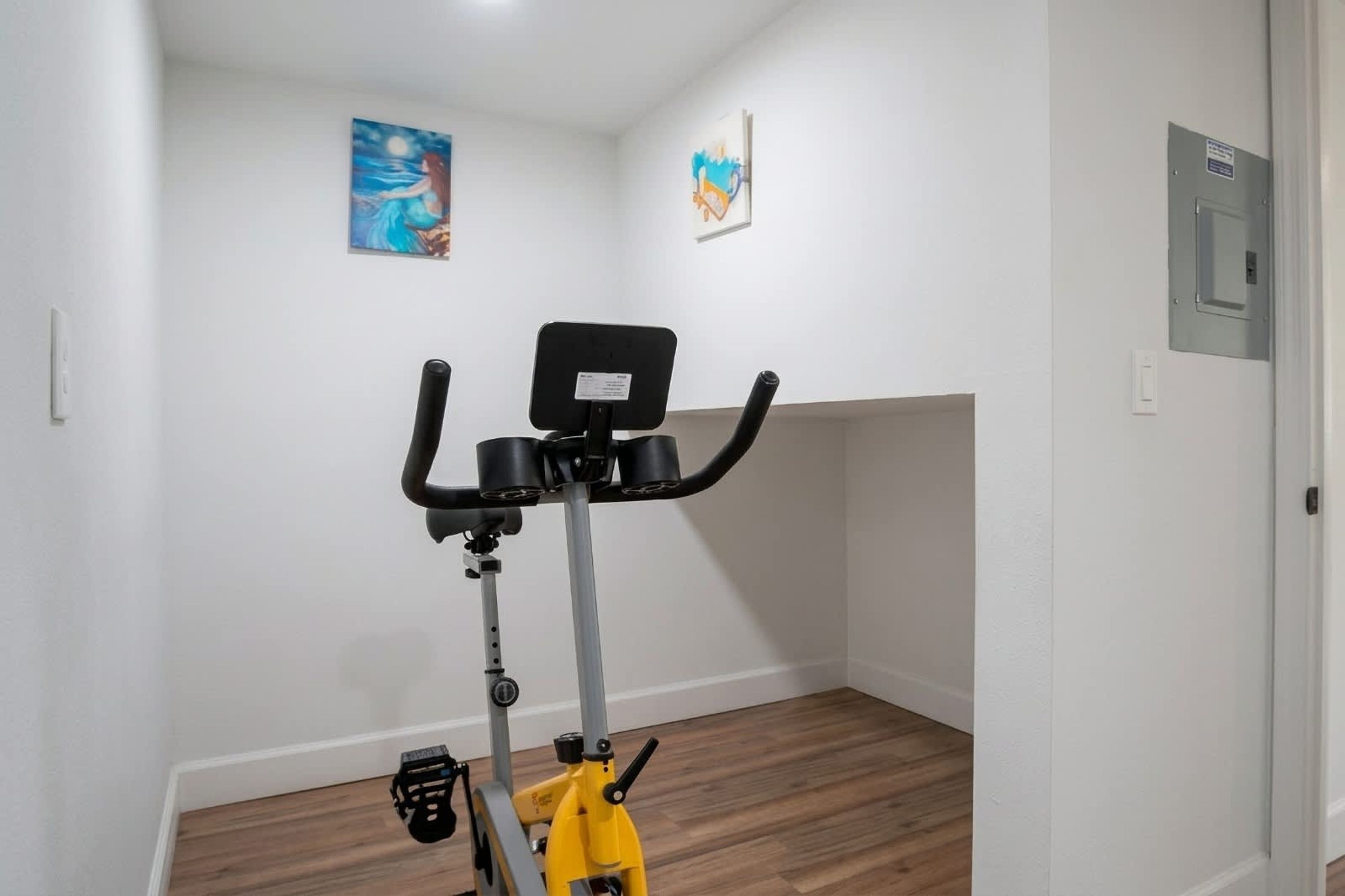 A room with a stationary bike, a door with a lock, and a wall-mounted electrical box.