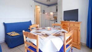 Ein gemütlicher Wohnraum mit Esstisch, Stühlen, einer Couch, einem Fernseher und einer kleinen Küchenzeile im Hintergrund.