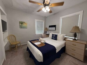 Quarto com cama, poltrona, ventilador de teto, janela com persianas, mesa de cabeceira e abajur.
