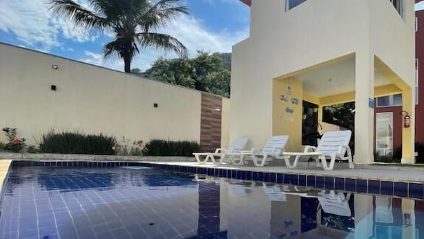 A swimming pool with tiled flooring, a palm tree, and a building with a yellow facade and white lounge chairs. - Residencial 220 Maresias (São Sebastião)