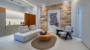 Ein modernes Wohnzimmer mit einer Couch, einem Couchtisch und Sesseln, einer Wand aus Stein als Gestaltungselement sowie einer Küchenzeile mit Schränken und Geräten.