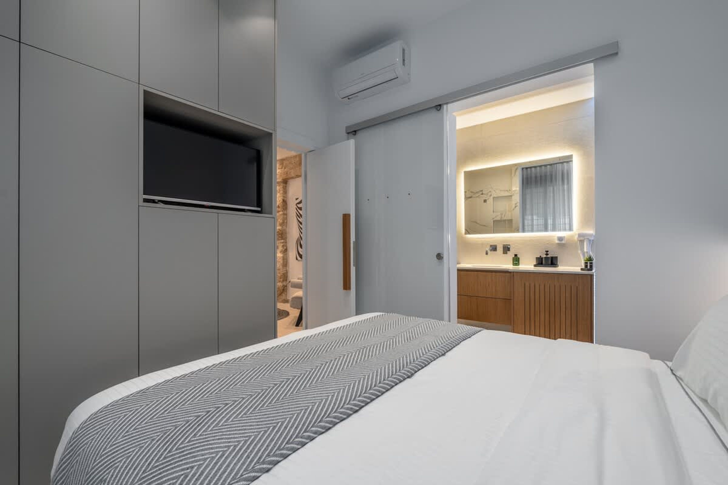 A modern hotel room with a bed, a TV mounted on the wall, a bathroom with a mirror, and a kitchenette.