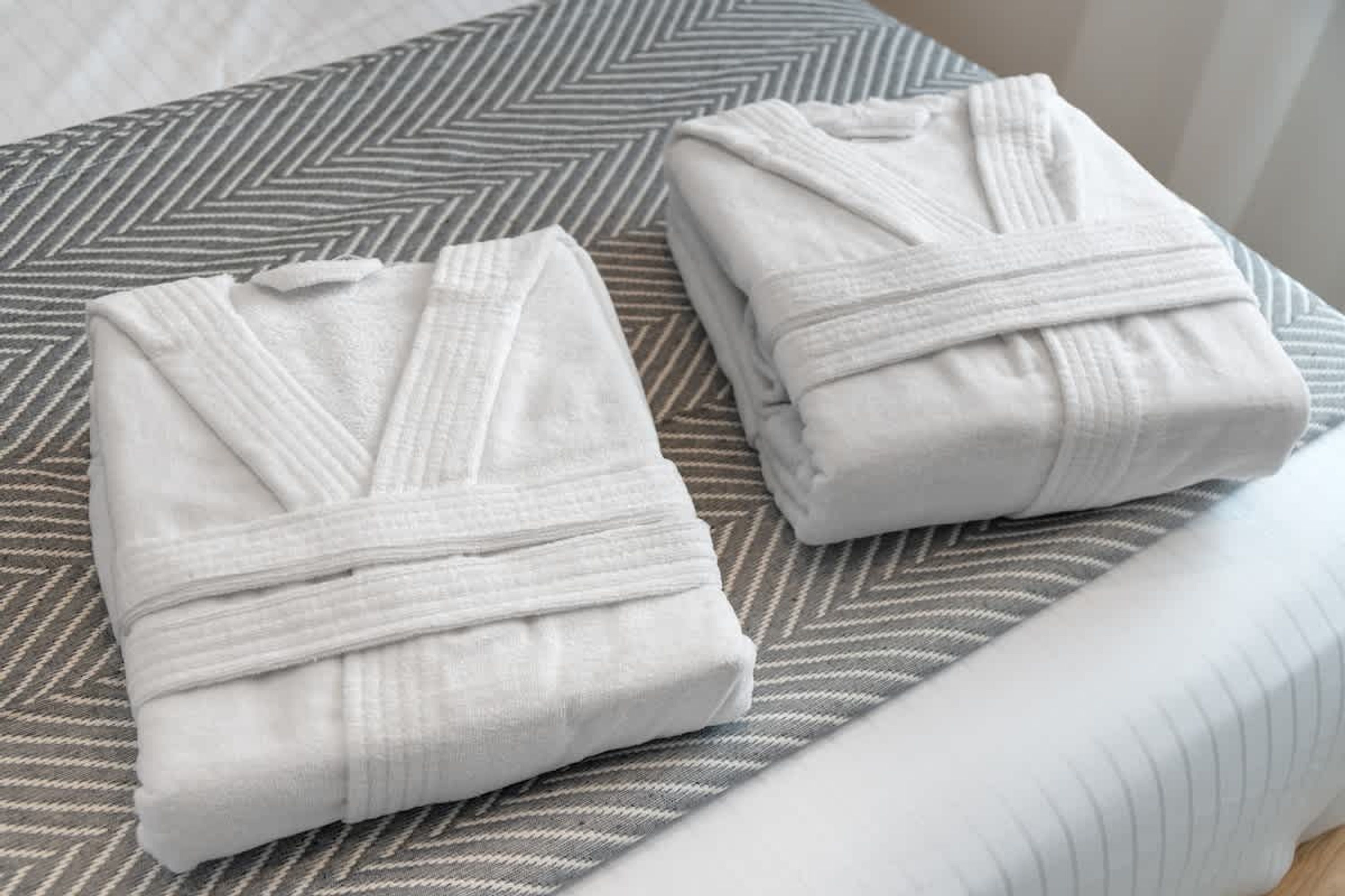 Two neatly folded white robes on a bed with a chevron-patterned pillow.