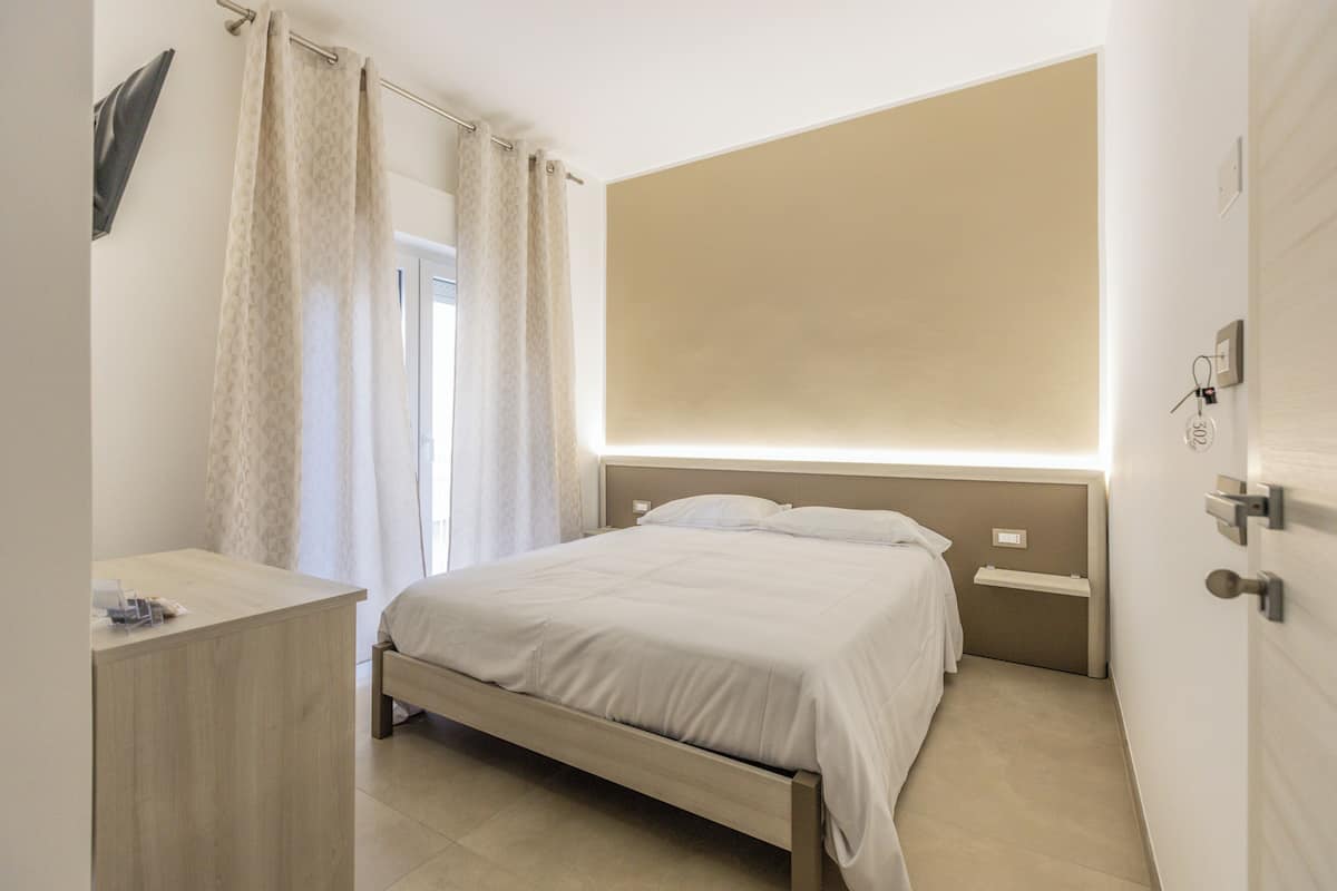 A bedroom with a bed, bedside tables, a desk, a window with curtains, and a wall-mounted light fixture.