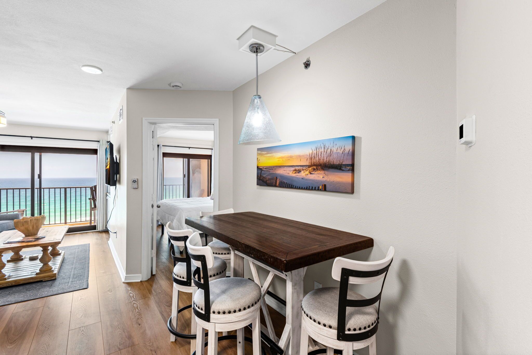 A dining area with a wooden table and white chairs, a pendant light, and a painting on the wall.