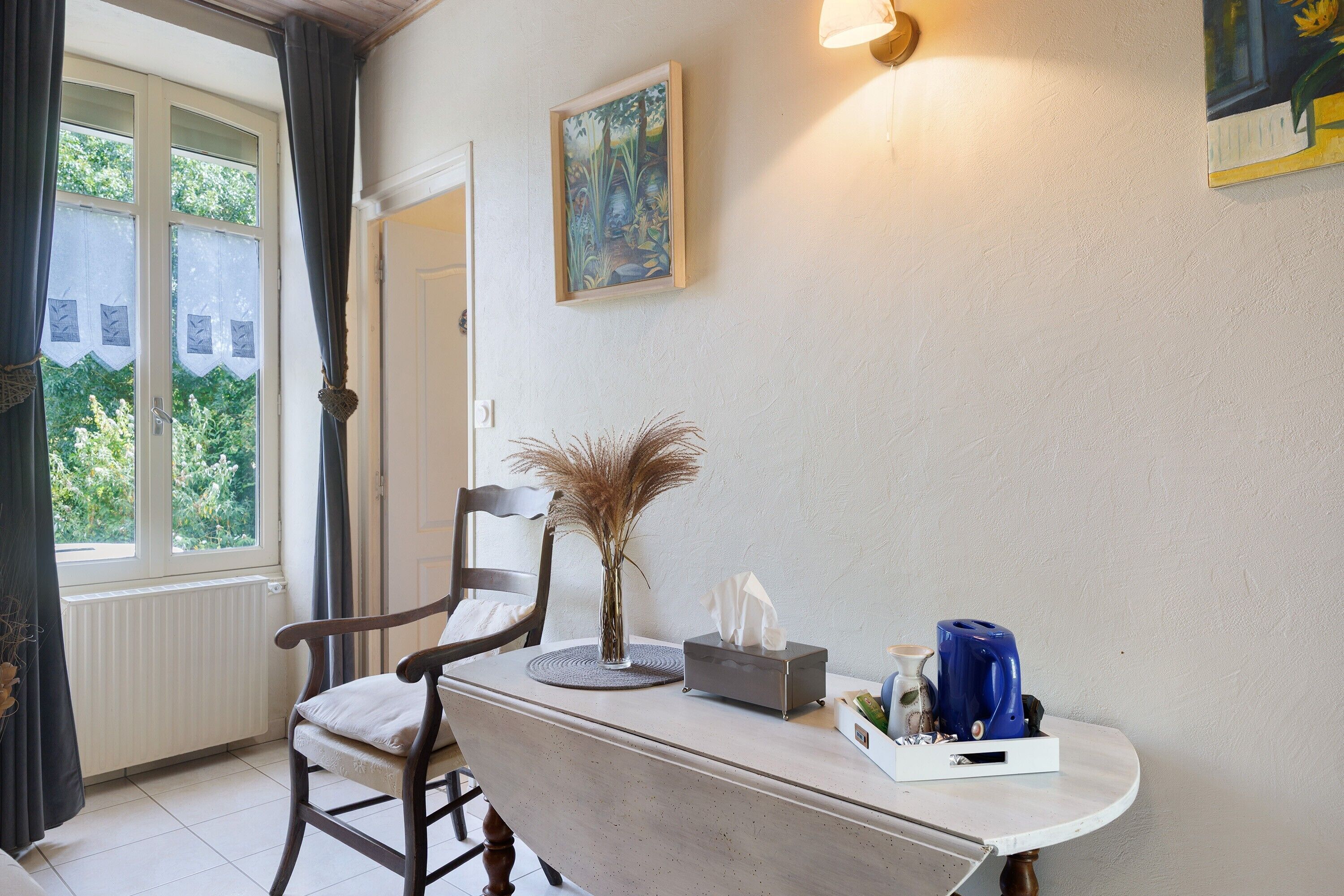 Une pièce comprenant une table à manger, une chaise, un vase contenant des plantes séchées, une boîte de mouchoirs, une bouilloire bleue et un tableau accroché au mur.