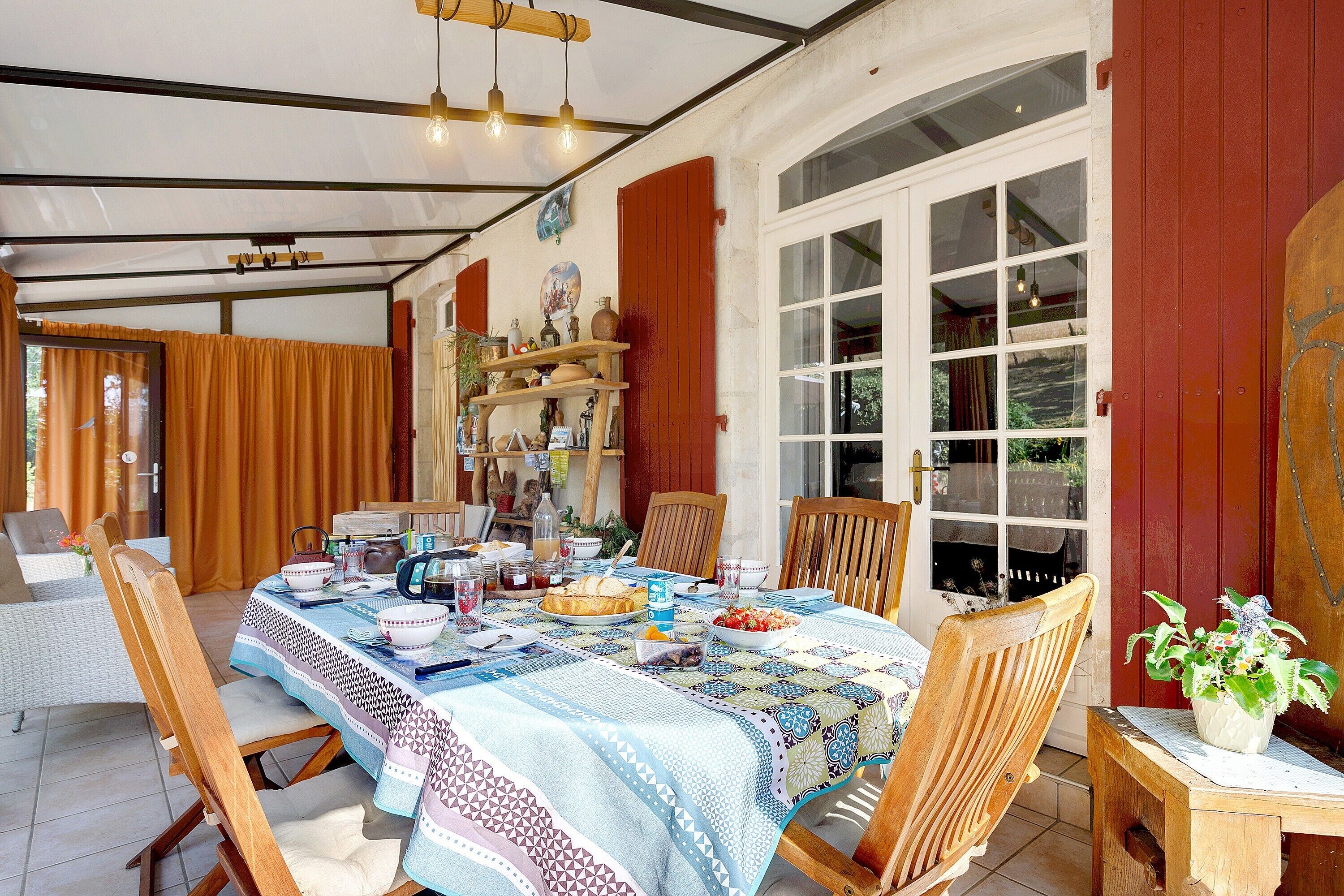 A dining area with a table set for a meal, chairs, a wooden sideboard with decorative items, and a large window with curtains.