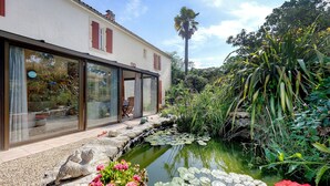 A house with a glass door leading to a patio, a small pond with lily pads, and a palm tree in the background.