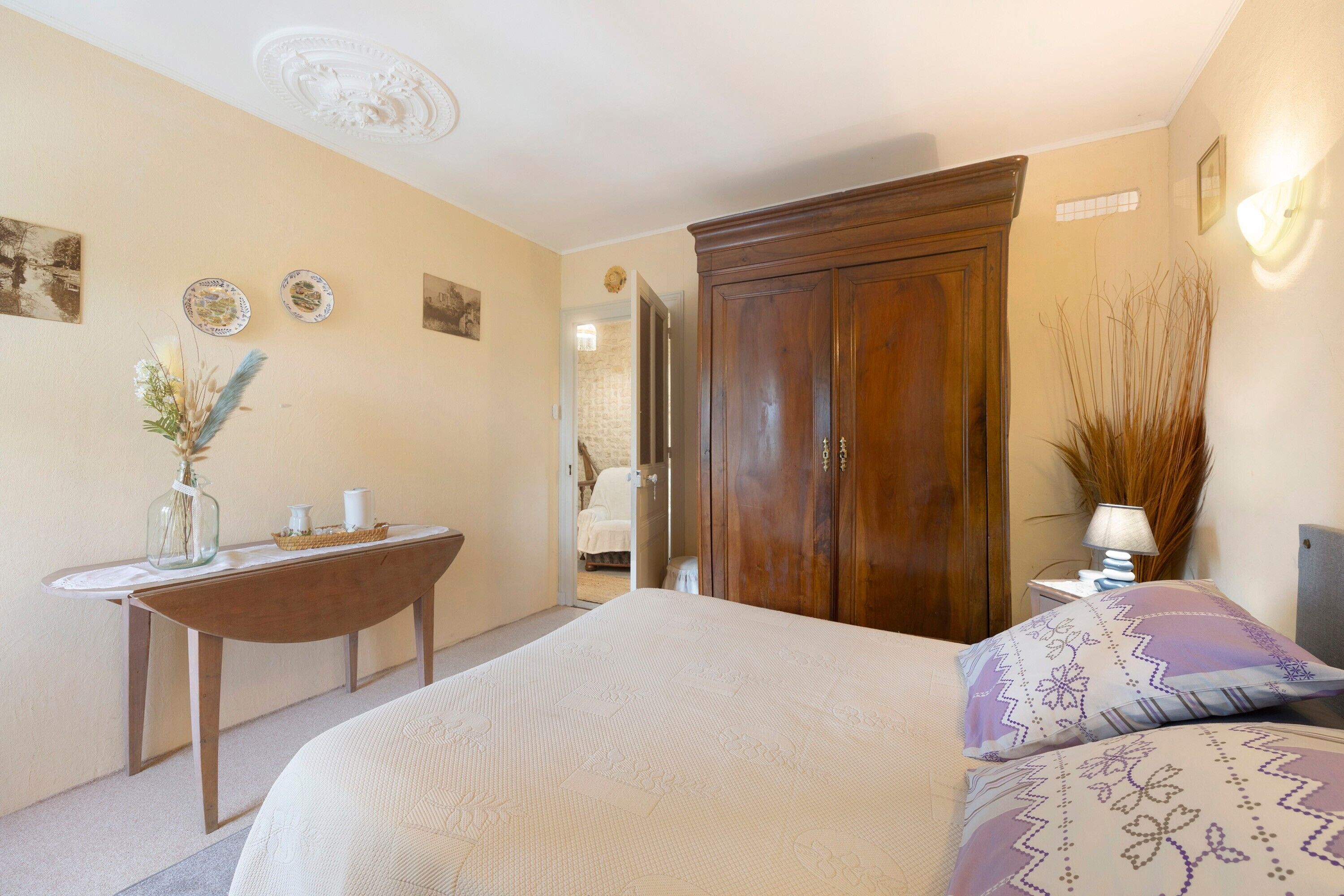 Un dormitorio con una cama, un armario de madera, una mesita de noche con una lámpara y una pequeña mesa con adornos.