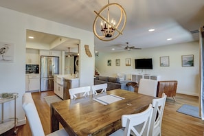 Eine moderne Küche mit Edelstahlgeräten, ein Essbereich mit Holztisch und weißen Stühlen sowie ein Wohnzimmer mit Sofa und Fernseher.