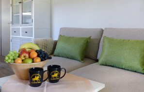Ein Wohnzimmer mit einer Couch, grünen Kissen, einem Holztisch mit Obst und zwei Tassen sowie einem weißen Schrank mit Glastüren.