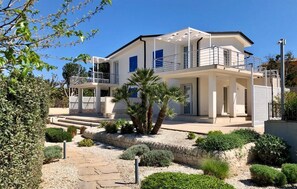 A modern two-story house with a balcony, a stone pathway, and various shrubs. - Cozy apartment in Reitani with swimming pool (Reitani)