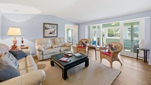 A living room with a sofa, armchair, coffee table, and a view through large windows. - Beaufort's Pink Paradise, Spacious home, steps away from Front Street (Beaufort)