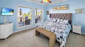 Un dormitorio con cama, ventilador de techo, televisor, ventana con vista a la playa y un cartel que dice 'HOME IS WHERE THE BEACH IS'.