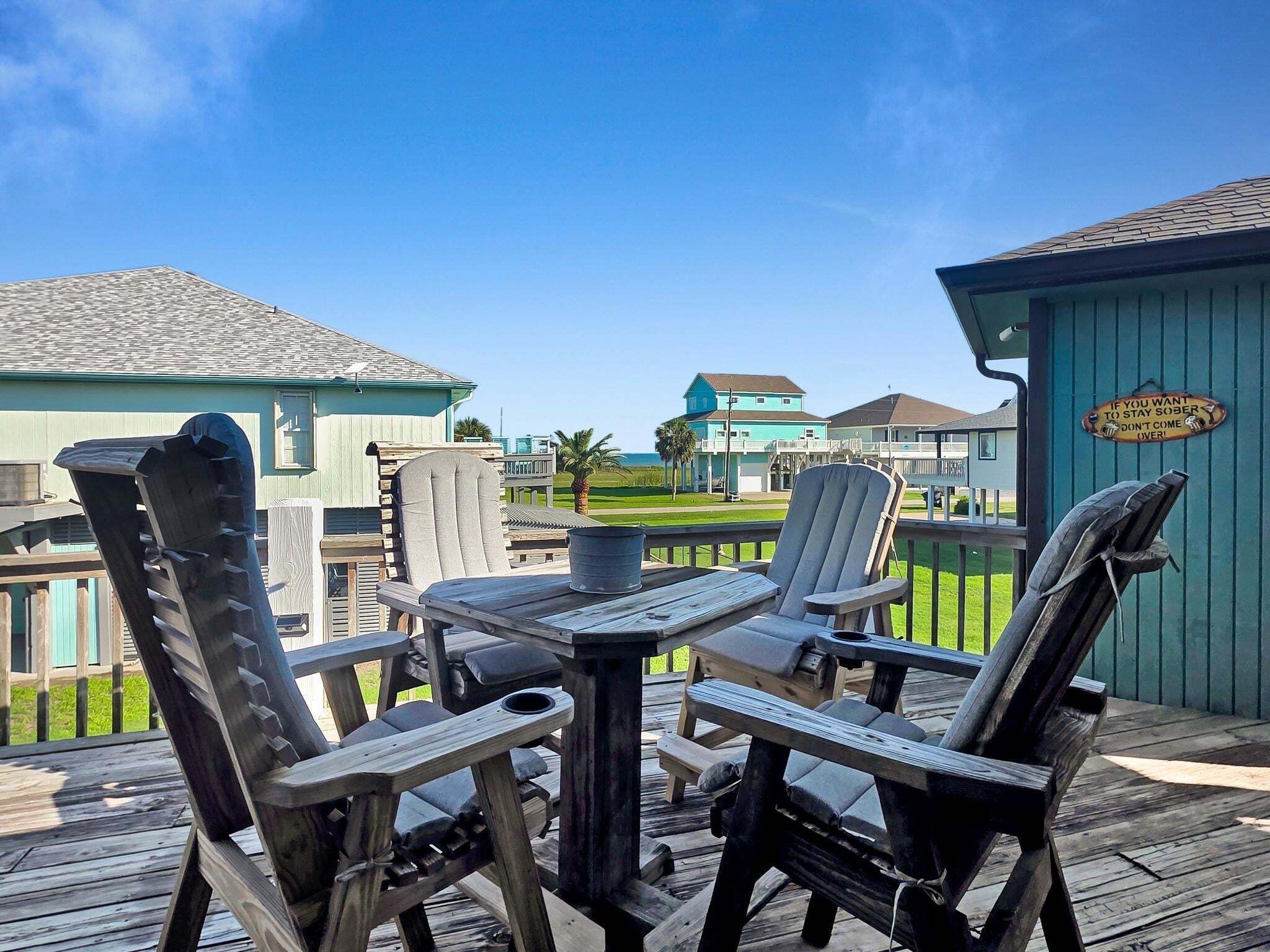 A wooden deck with Adirondack chairs, a table, and a view of buildings and a golf course.