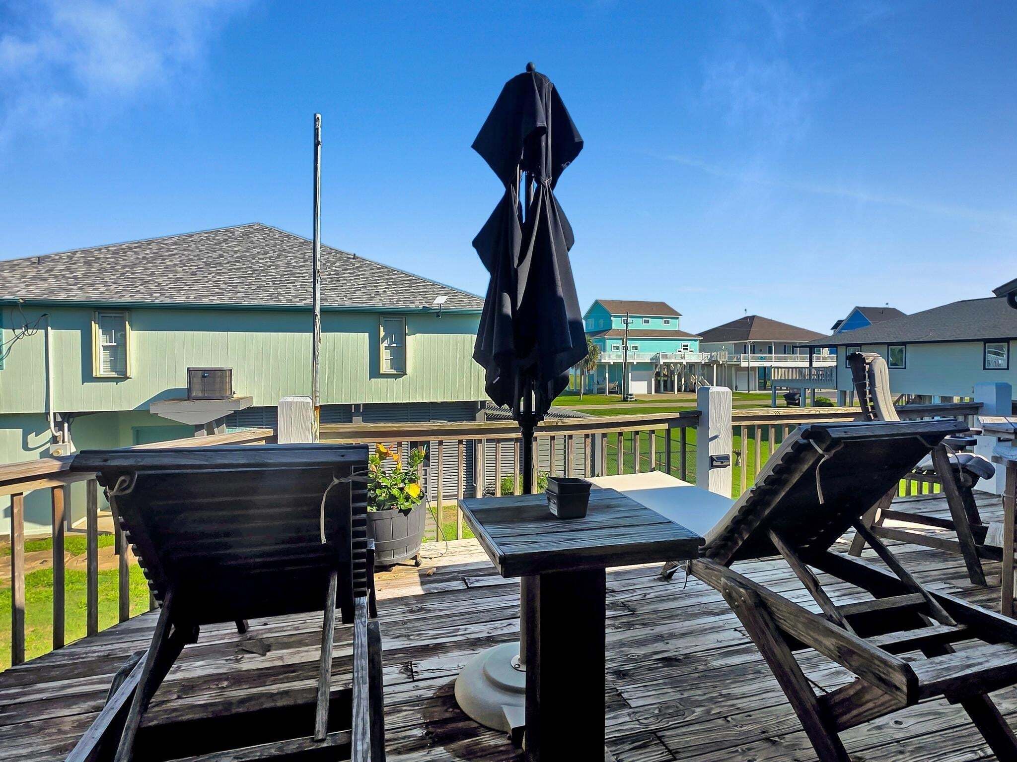 A wooden deck with a table and chairs, a black umbrella, and a view of houses in the background.