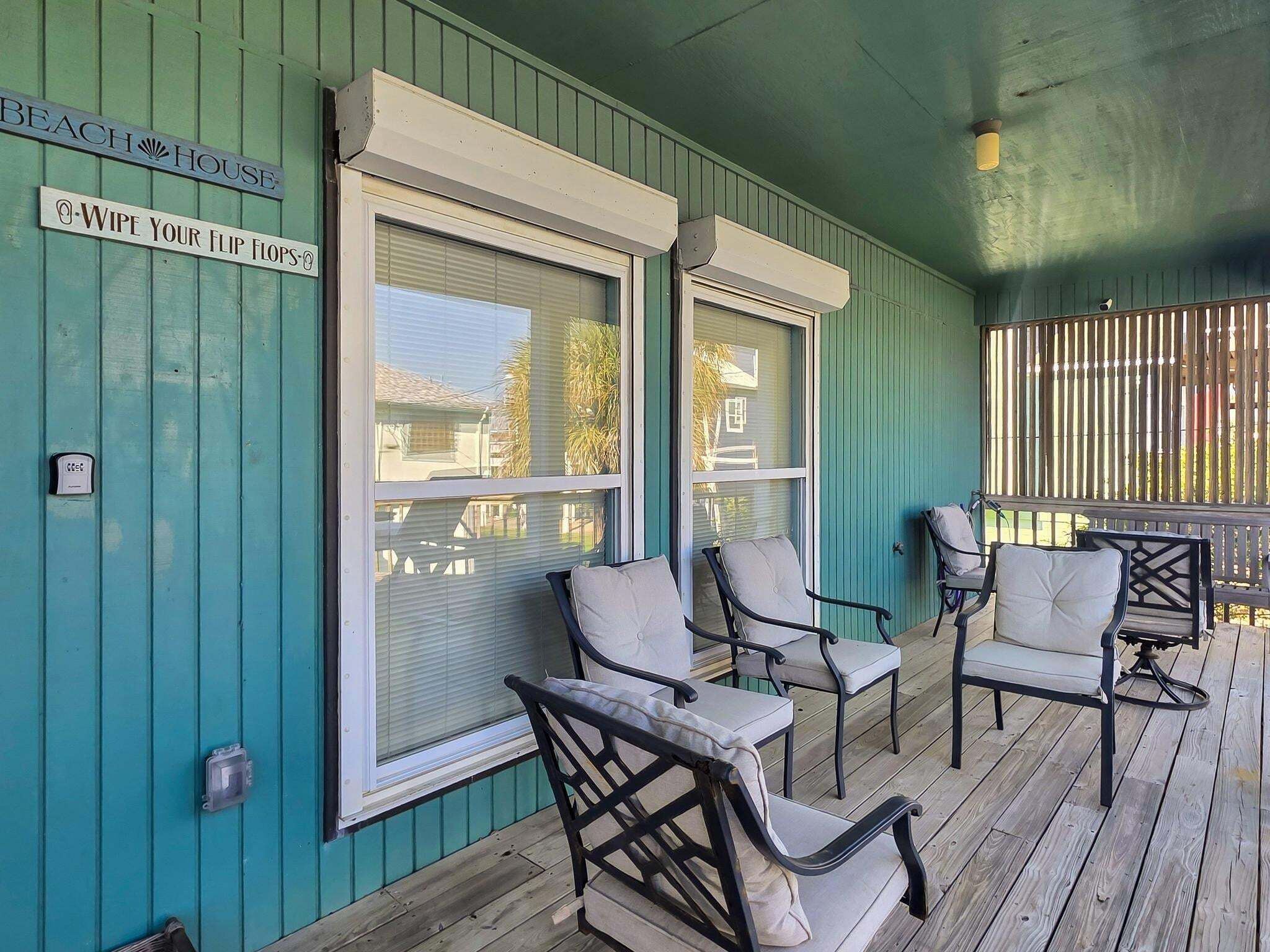 Een overdekt terras met buitenmeubelen, een schuifpui en een bordje met de tekst 'BEACH HOUSE'.