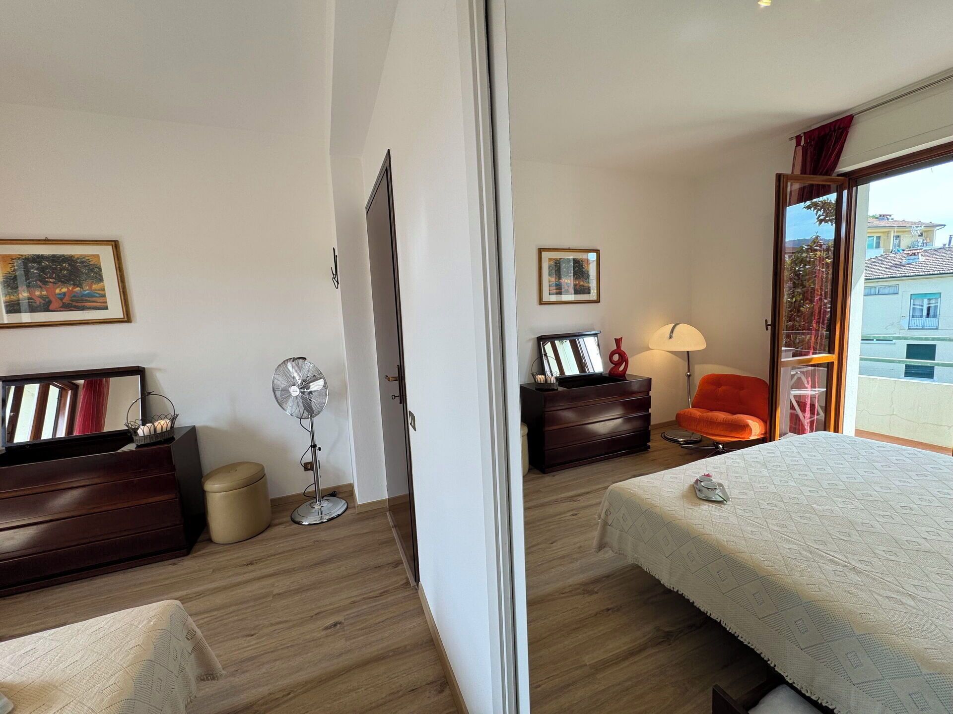 Ein Hotelzimmer mit Bett, Schreibtisch, Stuhl, Kommode, Stehlampe und einem Fenster mit Blick auf Gebäude.