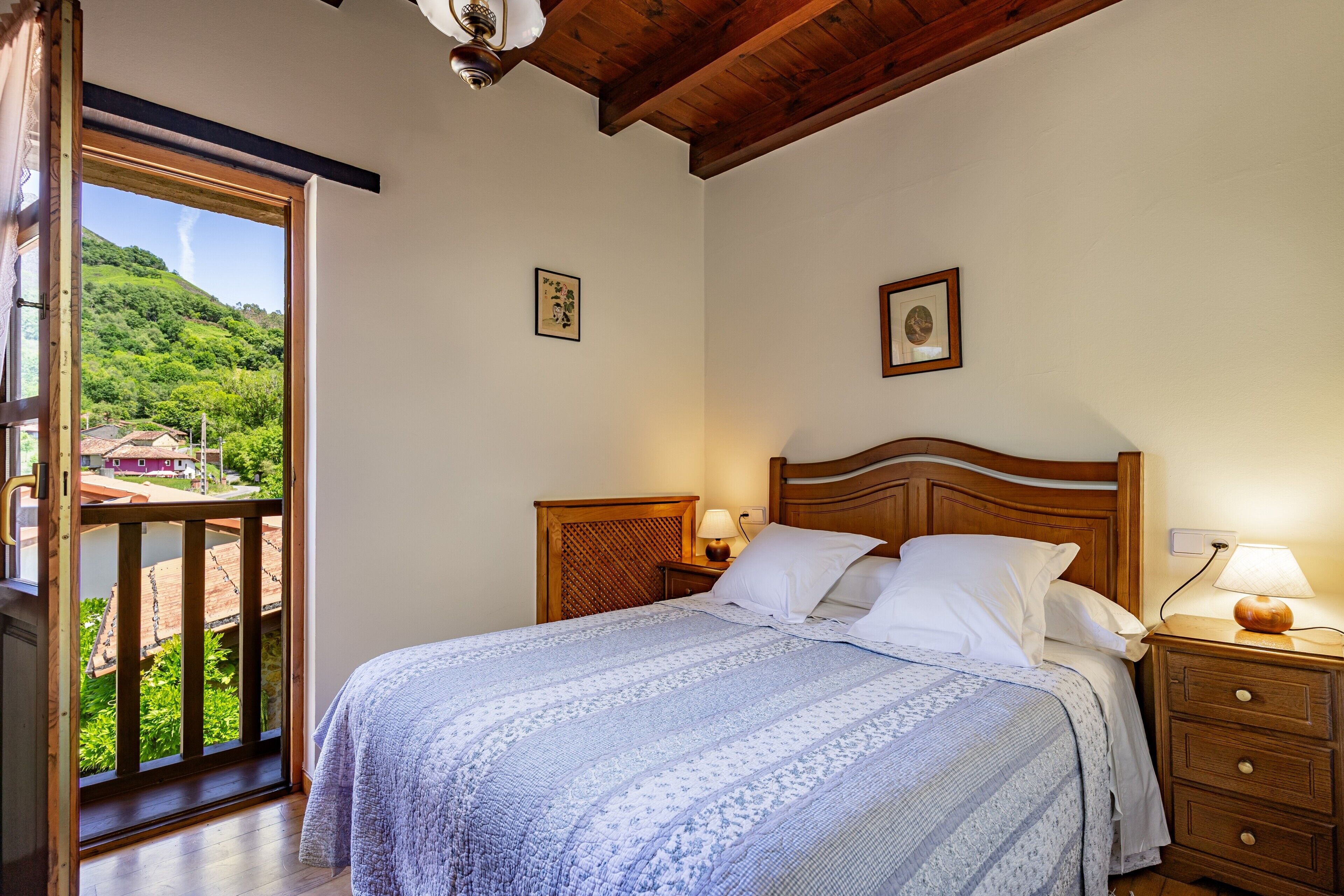 Un dormitorio con cama, cabecero de madera, mesitas de noche y vistas a un paisaje verde a través de un ventanal grande.