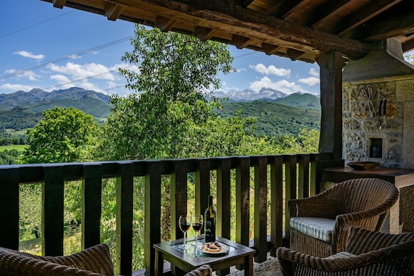 Un balcón de madera con muebles de mimbre, una mesa con una botella y vasos, y una vista panorámica de las montañas.