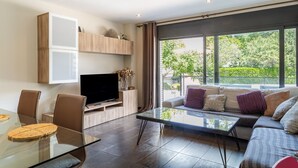 Ein modernes Wohnzimmer mit einem Glastisch, einer Couch mit Kissen, einem Flachbildfernseher und einem großen Fenster mit Blick ins Grüne.