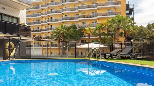 Ein Schwimmbecken mit einer Fliesenkante, umgeben von einem Zaun, und ein Gebäude mit Balkonen im Hintergrund.