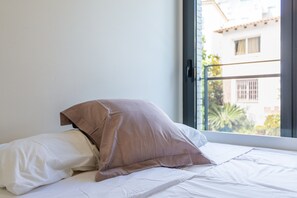 A bed with white bedding and a brown pillow, a window with a view of buildings and greenery, and a door with a handle. - Apartment 'Claret' with Mountain Views, Wi-Fi, and Air Conditioning (Lloret de Mar)