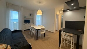 A modern living room with a sofa, dining table, and chairs, a TV, and a balcony with a view. - Sea View Escape at Lido dei Pini (San Michele al Tagliamento)