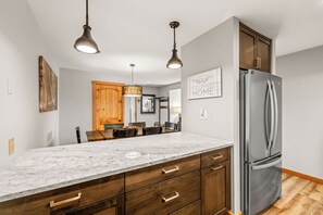 Eine Küche mit Marmorarbeitsplatte, Holzschränken, einem Edelstahlkühlschrank, Pendelleuchten und einem Essbereich mit Holztisch und Stühlen.