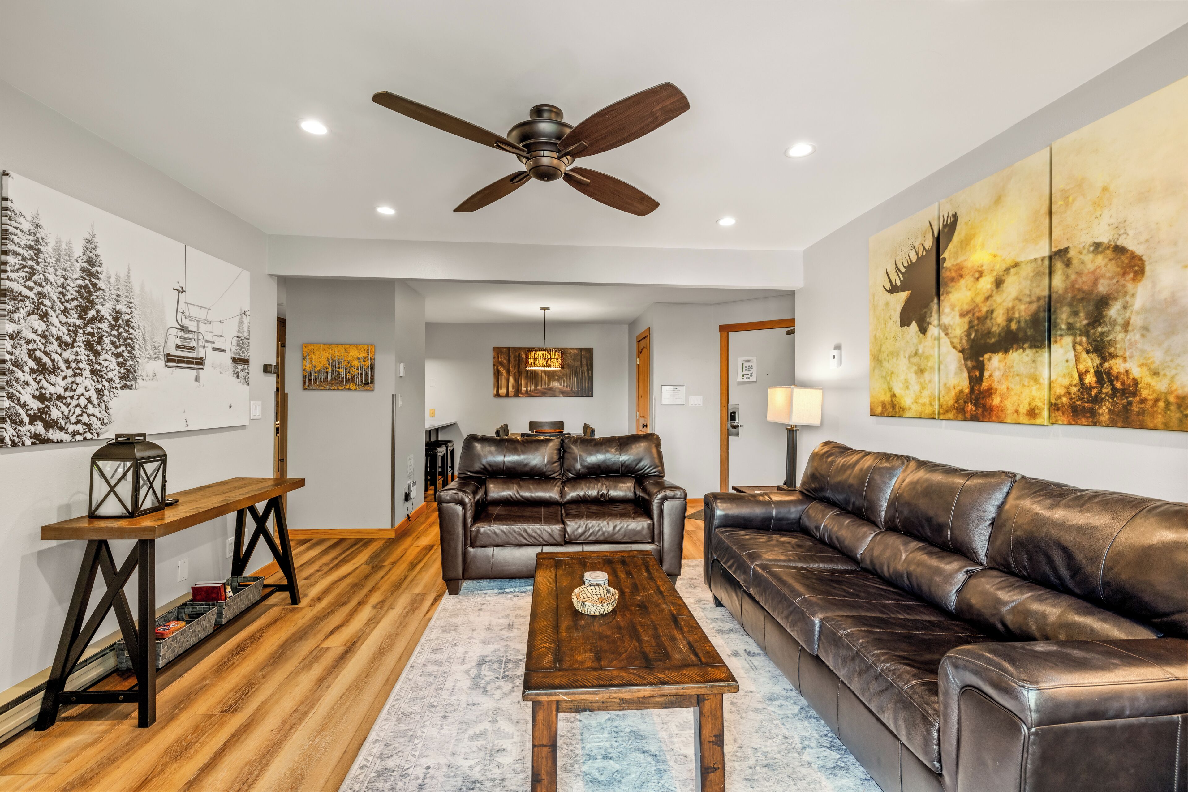 A living room with a brown leather sofa, a wooden coffee table, a ceiling fan, and artwork on the walls.