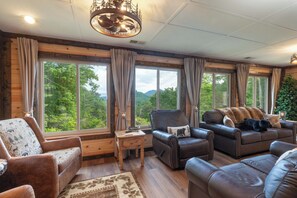 Ein gemütliches Wohnzimmer mit Ledersofas, Holzboden, großen Fenstern mit Vorhängen und einer Kristallleuchtre.