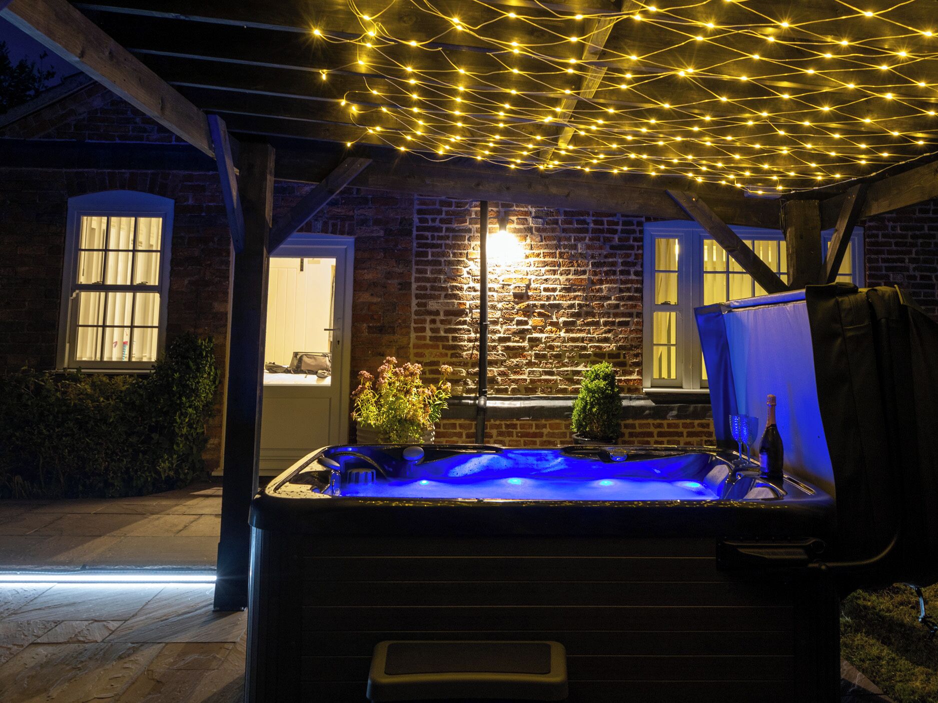 Un jacuzzi con luz azul, rodeado por un muro de ladrillo y vigas de madera, con luces colgantes en el techo.