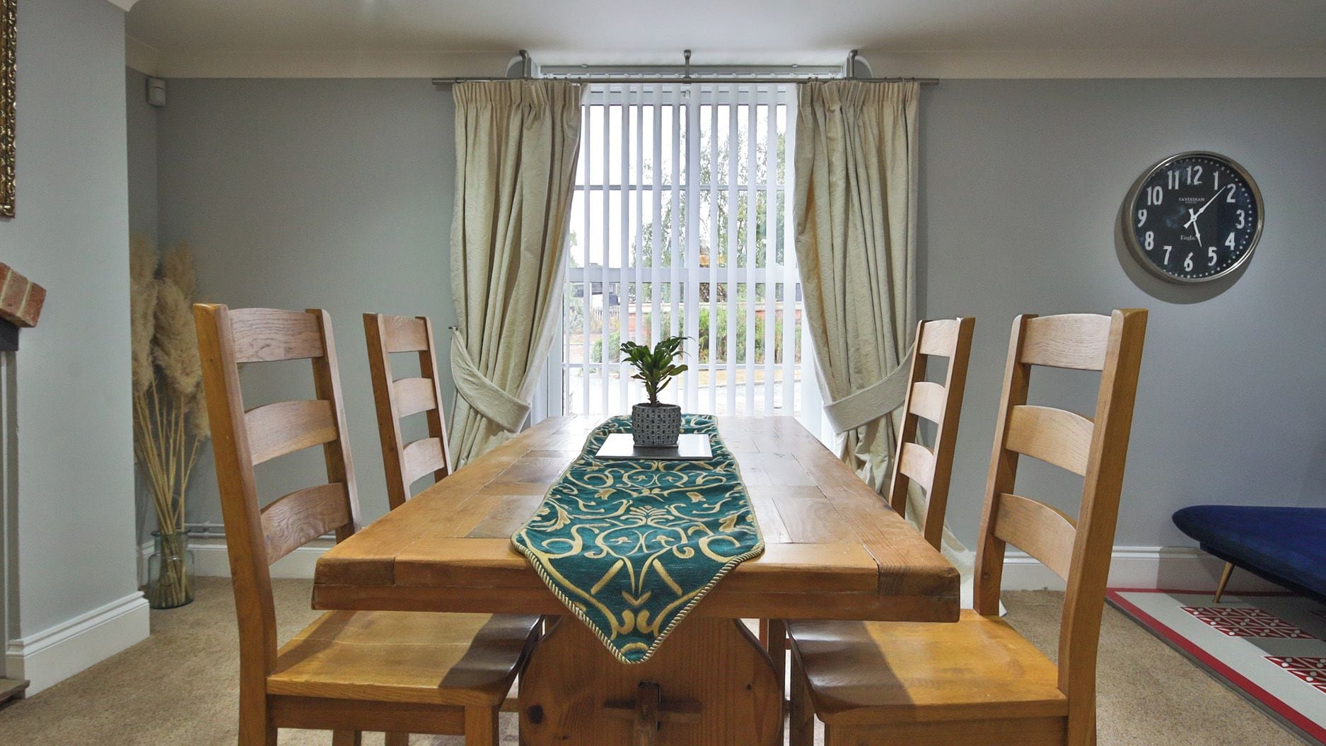 Un comedor con mesa de madera, sillas, un reloj y vistas a través de una ventana con cortinas.