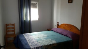 A bedroom with a bed, a wooden chair, a window with curtains, and a wall-mounted light switch. - Casa Olivo – Conil de la Frontera, 2 bedrooms, apartment, 4 guests (Conil de la Frontera)