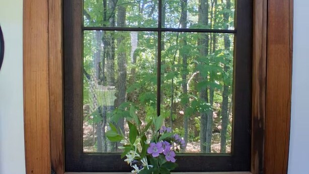 A window with a wooden frame and a mesh screen, a vase with flowers on a wooden ledge below, and a label on the window. - Beautiful custom tiny home 20 min from Middlebury! (Addison)