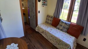 A room with a sofa bed, a wooden floor, a door leading to another room, and a window with curtains. - Beautiful custom tiny home 20 min from Middlebury! (Addison)