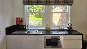 A kitchen with a black countertop, white cabinets, a stainless steel sink, a kettle, and a fire blanket on the wall.