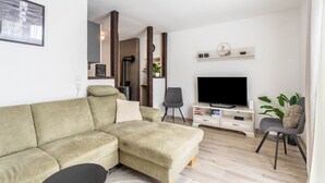 A modern living room with a sectional sofa, a flat-screen TV, a small white cabinet, and a grey chair. - 'Meini' Holiday Apartment in the Old Train Station (Brilon)