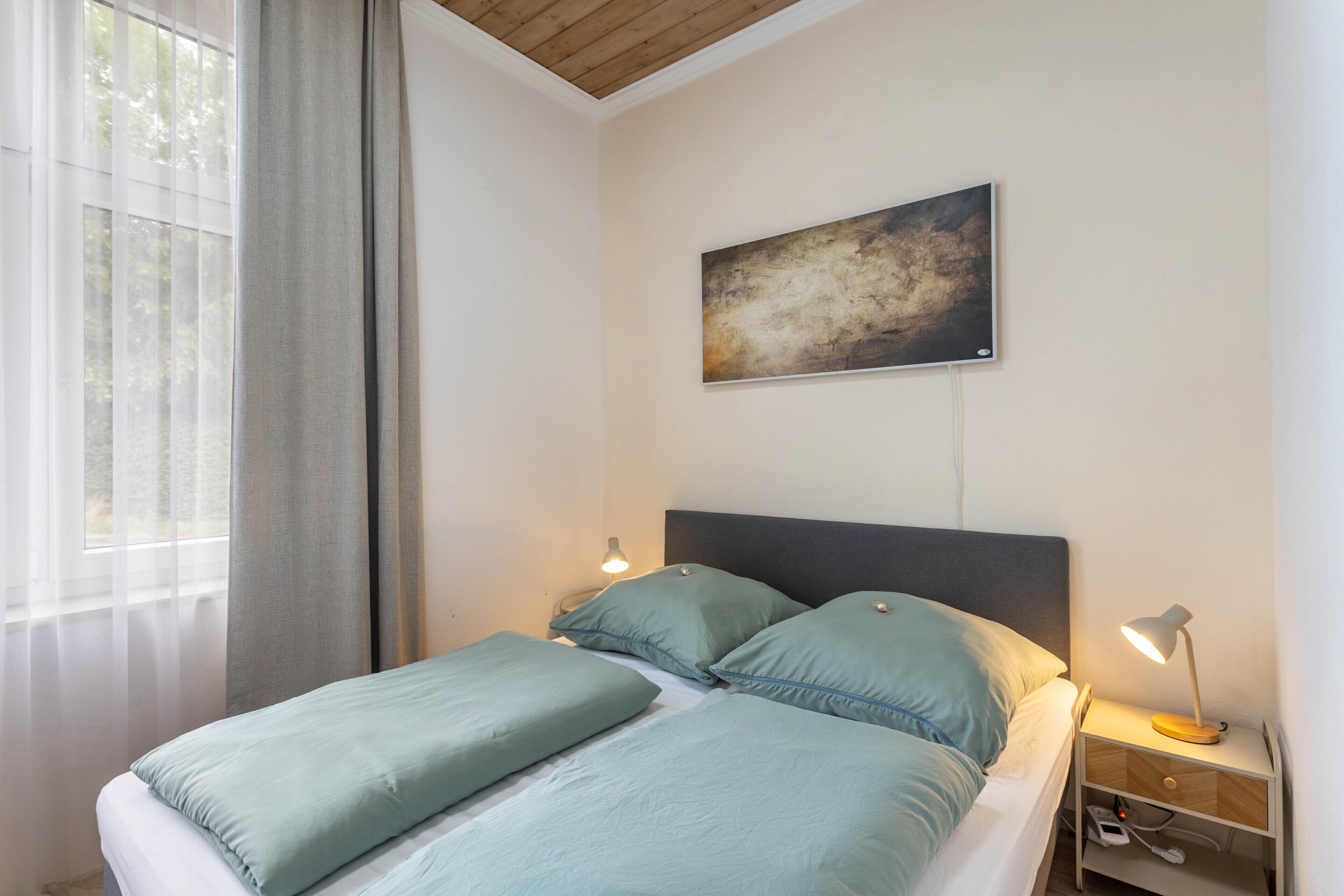 Ein Schlafzimmer mit Bett, Nachttisch, Lampe, einem Gemälde an der Wand und einem Fenster mit Vorhängen.