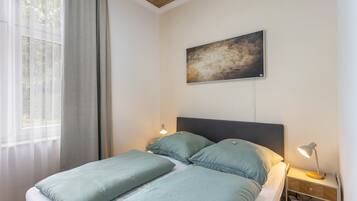 Ein Schlafzimmer mit Bett, Nachttisch, Lampe, einem Gemälde an der Wand und einem Fenster mit Vorhängen.