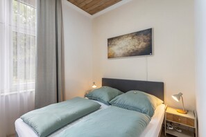 Un dormitorio con cama, mesita de noche, lámpara, un cuadro en la pared y ventana con cortinas.