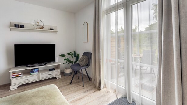 A modern living room with a flat-screen TV, a sofa, a chair, a plant, and a large window with curtains. - 'Meini' Holiday Apartment in the Old Train Station (Brilon)