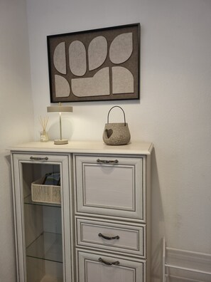 A white cabinet with glass doors and drawers, a framed abstract artwork above it, and a small table with a lamp and decorative items. - 'Meini' Holiday Apartment in the Old Train Station (Brilon)